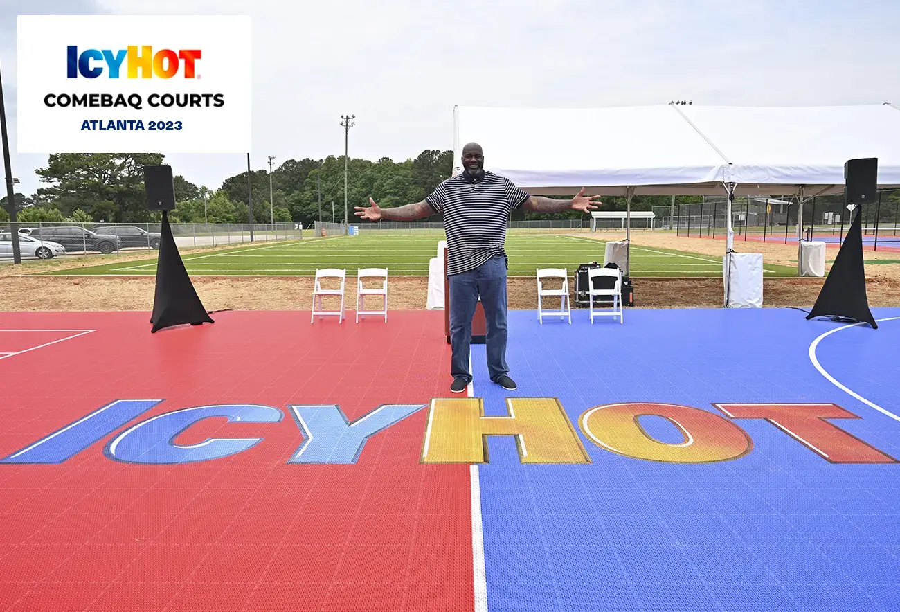 Shaquille O'Neal standing at the center of a newly renovated basketball court during the 2023 Icy Hot Comebaq Courts event in Atlanta. The basketball legend stands with open arms, exuding enthusiasm, atop the court that showcases a vibrant red and blue Icy Hot logo. In the upper-left corner of the image, a label displays the Icy Hot Comebaq Courts logo, along with the event's year (2023) and location (Atlanta).
