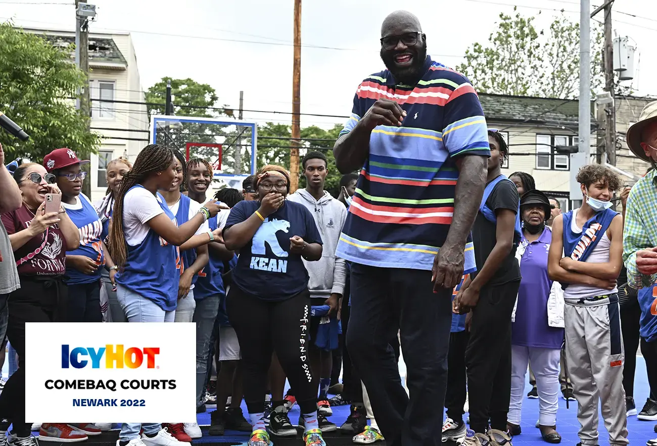 Shaquille O'Neal, wearing a warm smile, stands at the center of a newly renovated basketball court in Newark, surrounded by a group of enthusiastic young people. The lower-left corner of the image features a label displaying the Icy Hot Comebaq Courts logo, along with the event's year (2022) and location (Newark).