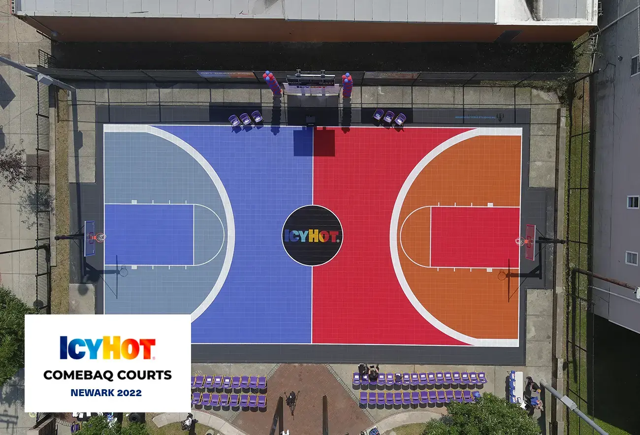 An impressive aerial view of the newly renovated basketball court in Newark, creatively divided with vibrant red and blue colors, representing the iconic Icy Hot logo. The court's centerpiece proudly displays the Icy Hot logo. The lower-left corner of the image features a label displaying the Icy Hot Comebaq Courts logo, along with the event's year (2022) and location (Newark).