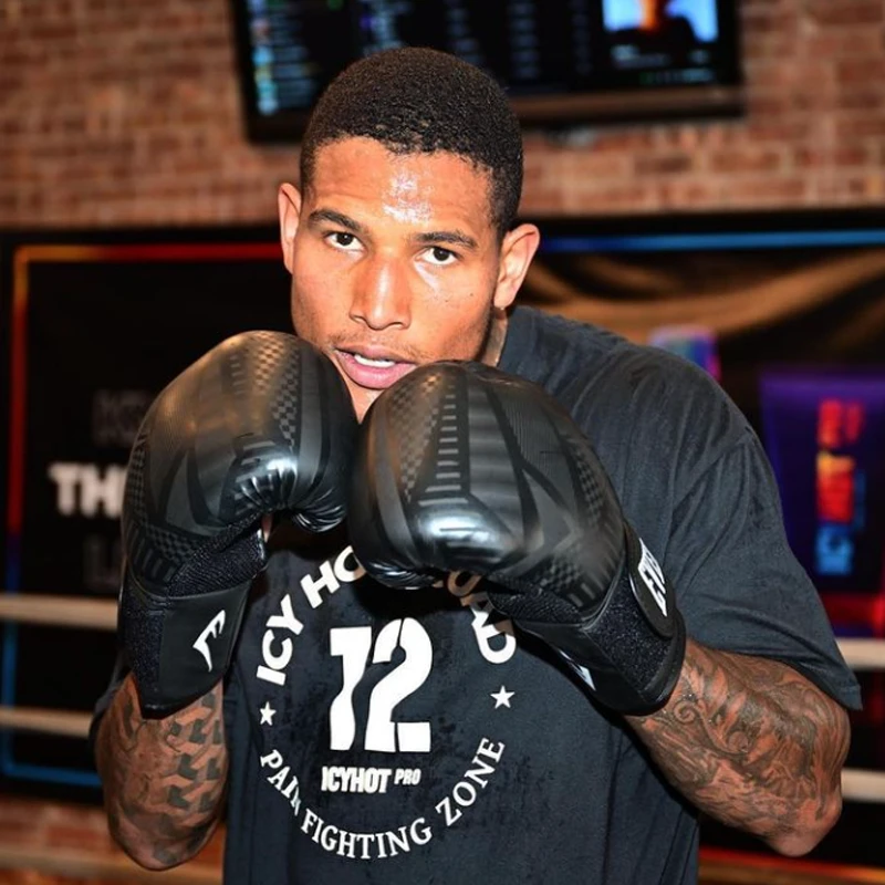 A close-up of Darren Cross facing the camera, standing on a boxing ring, in guard pose. He wears boxing gloves, and a t-shirt that reads "Icy Hot Squad - 72 - Icy Hot PRO - Pain Fighting Zone".